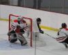 Wesleyan's goalie in White red and black with a red logo stands in net as a Wesleyan player skates behind the net to confront a Salve Regina player in black or navy.